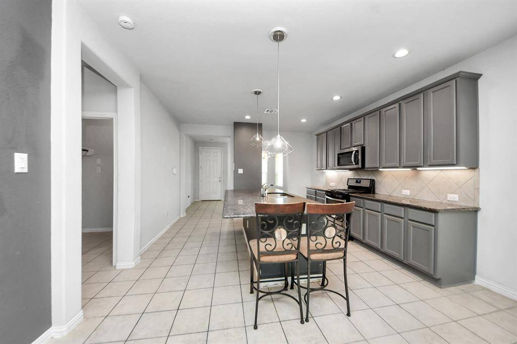 This modern kitchen features sleek gray cabinets, granite countertops, and stainless steel appliances. An island with bar seating and pendant lighting adds functionality and style. The space is bright with recessed lighting and a tiled floor.
