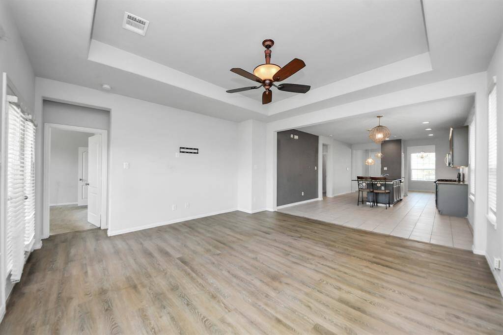This bright, open-concept living area features modern wood flooring, a ceiling fan, and ample natural light from multiple windows. It seamlessly connects to the tiled kitchen, offering a spacious and inviting atmosphere.