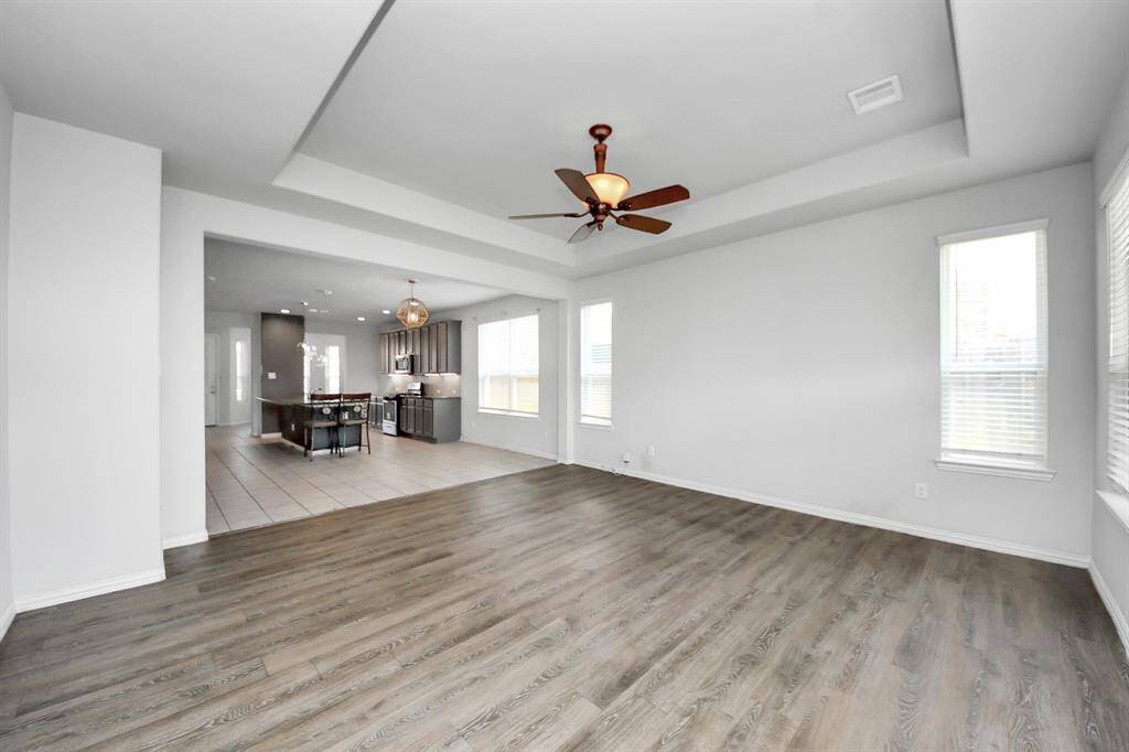 This spacious, bright room features light wood flooring, a tray ceiling with a ceiling fan, and plenty of natural light from large windows and glass patio doors.