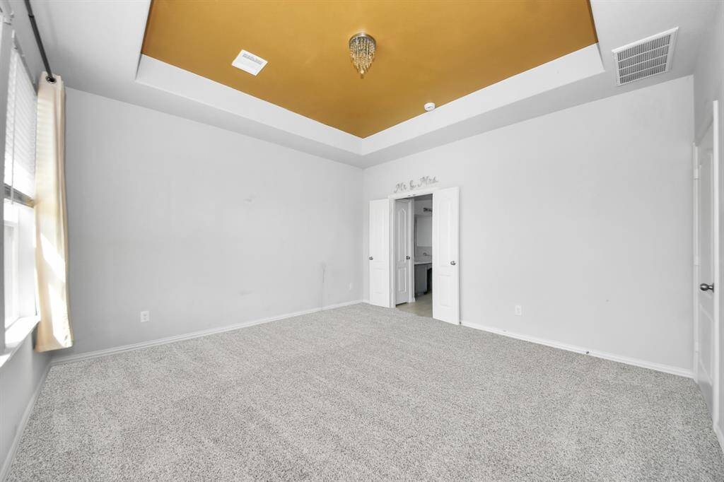 Spacious room with soft gray walls, plush carpet, and a striking gold tray ceiling with a chandelier. Features a large window for natural light and double doors leading to an adjacent area.