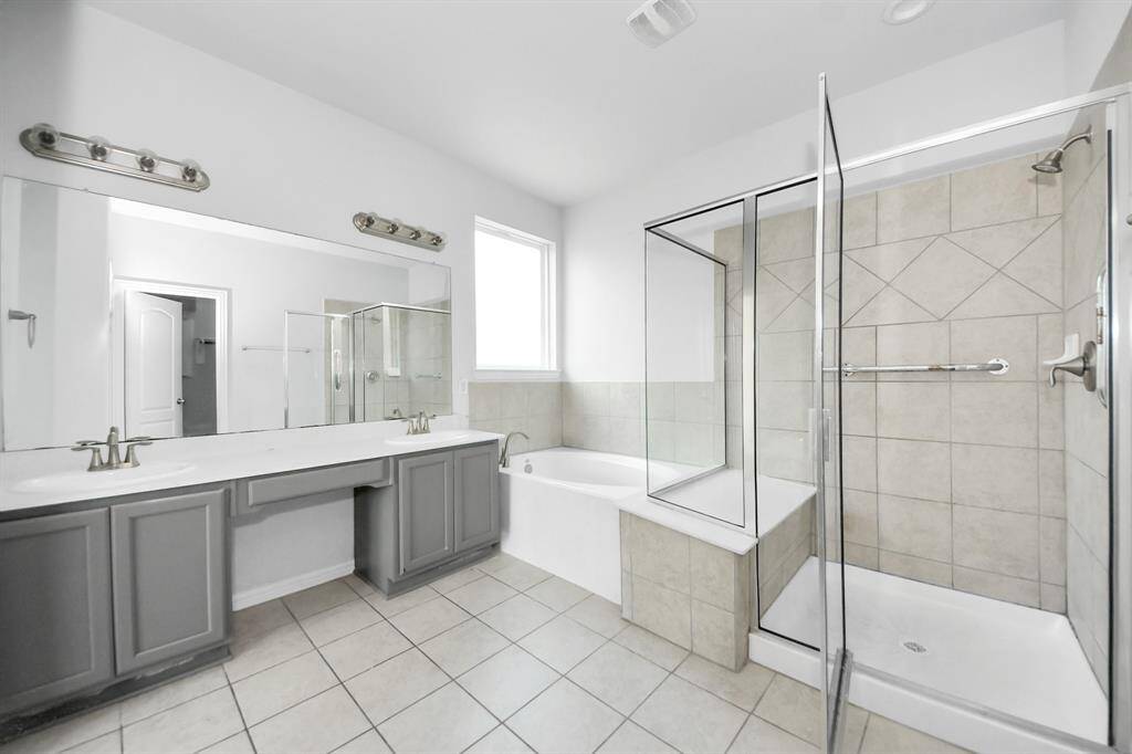 This spacious bathroom features a dual-sink vanity with ample storage, a large mirror, a walk-in shower, and a separate bathtub. The neutral tile flooring and walls create a clean, modern look.