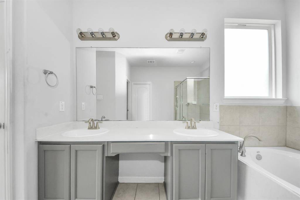 This bathroom features a double sink vanity with modern fixtures, a large mirror, and a nearby bathtub. The space is bright with natural light from a window, complemented by neutral tones for a clean, contemporary look.