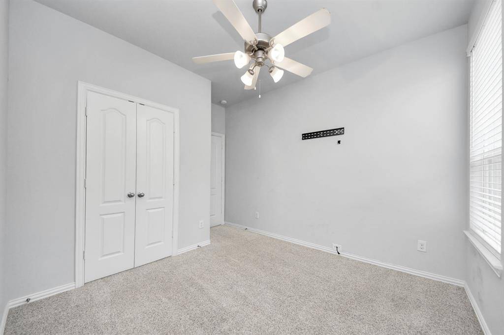 This room features soft gray walls, a ceiling fan with lights, carpeting, a double-door closet, and natural light from a large window, offering a cozy and functional space.