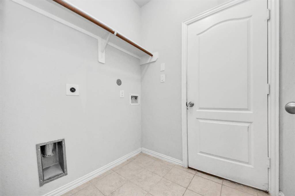 This photo shows a laundry room with hookups for a washer and dryer, a shelf for storage, and a white door. The room has neutral-colored walls and tile flooring.