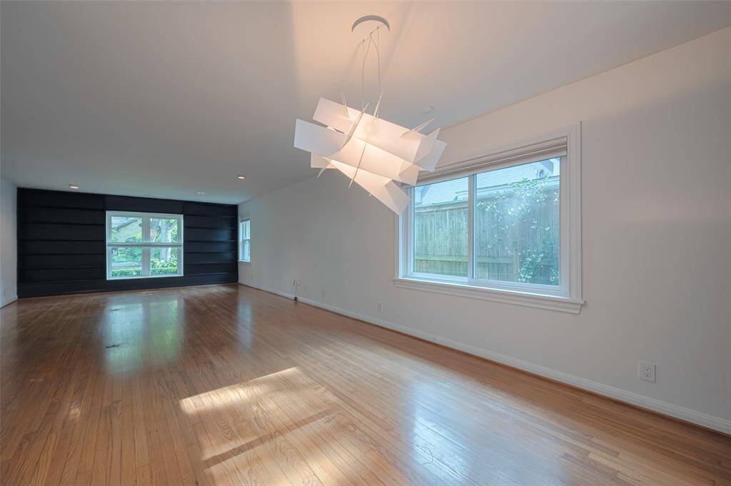 This photo showcase living and dining room with hardwood floors, a large window with abundant natural light, and a modern chandelier. The dark accent wall at the far end offers a stylish contrast, enhancing the room's contemporary feel.
