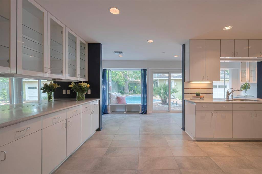 This bright kitchen features ample cabinetry with glass doors, modern tile flooring, and a spacious layout. Large windows and sliding doors offer a view of the backyard pool, bringing in natural light and a serene atmosphere.