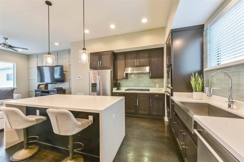 This modern kitchen featuressleek quartz countertops, richdark cabinetry, and a stylishsubway tile backsplash. A largewaterfall island offers ampleseating and workspace,complemented by chic pendantlighting. Stainless steelappliances, including a gascooktop and built-in microwave,add to the contemporary appeal.A deep farmhouse sink beneatha bright window enhancesfunctionality, while recessedlighting and an open layout makethis space perfect for cookingand entertaining.