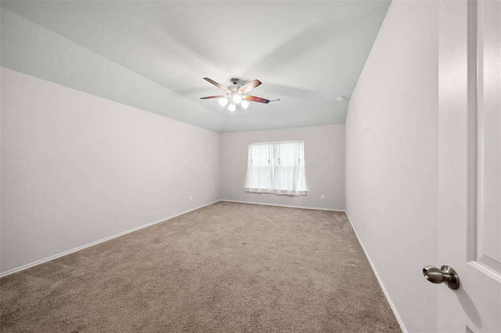 This is a bright and spacious room with wood flooring, neutral walls, and ample natural light. It features a ceiling fan and large windows overlooking a green outdoor area, offering a cozy and inviting atmosphere.