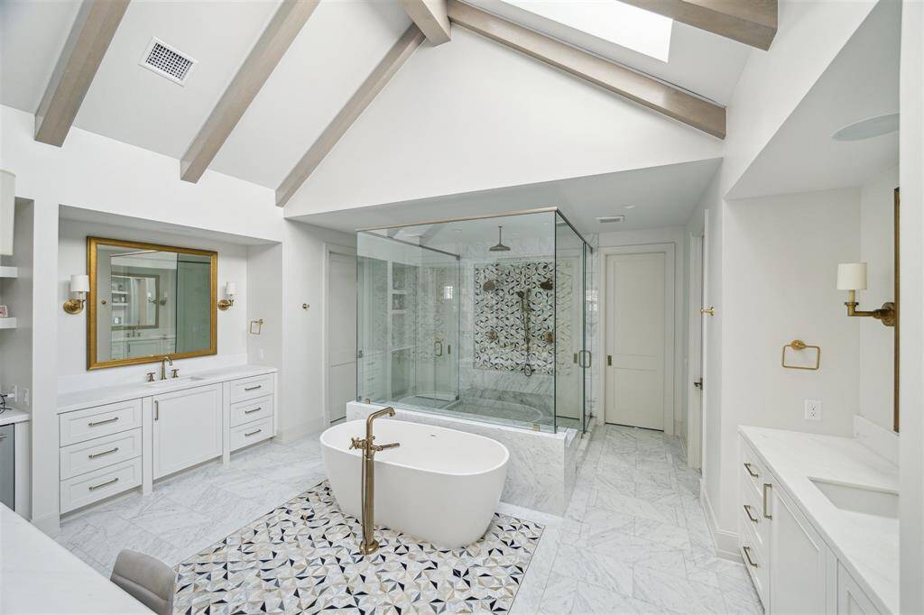 The serene primary bath is a true oasis, featuring majestic white pine ceiling beams with LED trim details that enhance its tranquil ambiance. Skylights flood the vaulted ceiling with natural light, further elevating the sense of relaxation and luxury. Custom cabinetry, separate vanities with luxe gold hardware compliment ample space for toiletries and more.