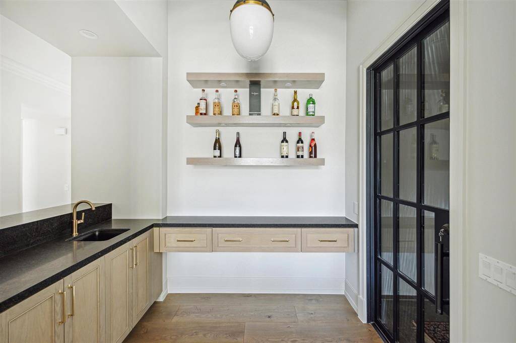 Perfectly positioned off the formal living room, this exquisite wet bar features sleek Black Misted honed quartzite countertops, white oak built-in shelves with art lighting, and direct access to the walk-in climate-controlled wine room, creating an ideal space for entertaining.