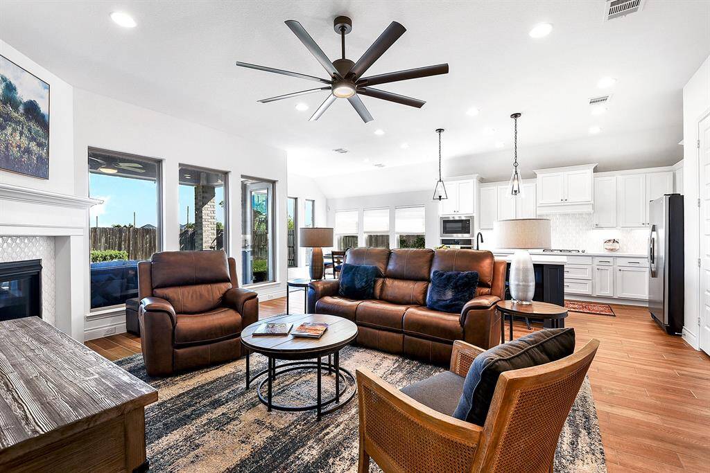 The living room is open to the kitchen and dining room, featuring high ceilings, a white-tiled gas fireplace with a mantel, wood-like tiled floors, recessed lighting, and floor-to-ceiling windows.