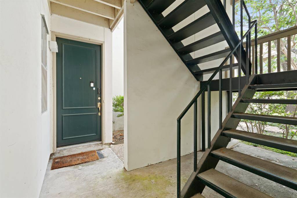 Lush greenery surrounds the entryway, with neatly trimmed bushes and mature trees adding a natural, refreshing element to the space. The staircase sits off to the side, providing a convenient path to the upper-level units for neighboring residents