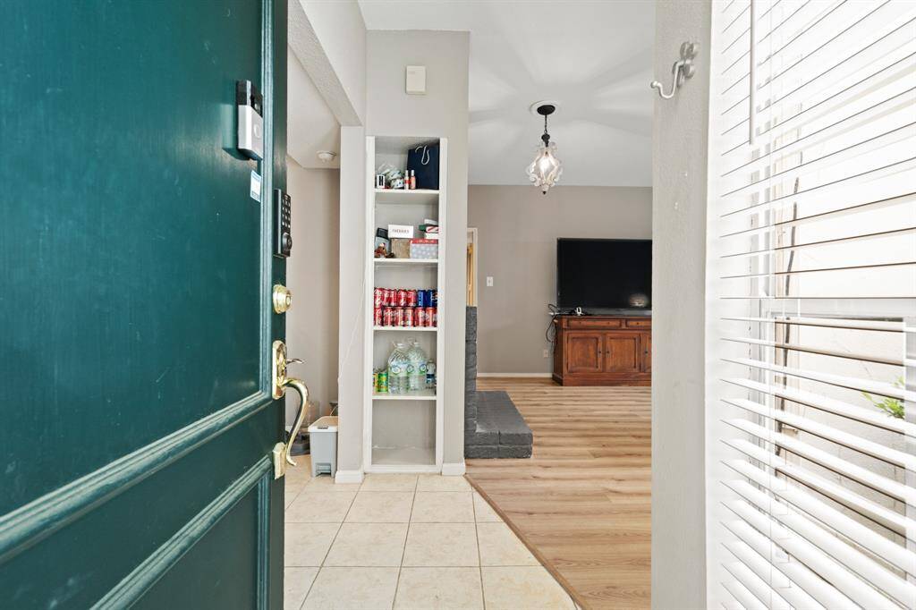 As you step inside, you’re greeted by a bright, open living area, that creates a warm and inviting atmosphere. Natural light filters through the window blinds to the right, complementing the contemporary light fixture overhead for a balanced and airy feel.