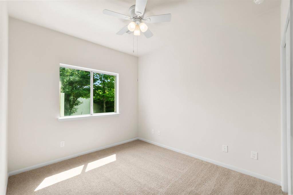 Bright, cozy bedroom with neutral walls, a ceiling fan, and a window overlooking greenery. Carpeted floor adds warmth.