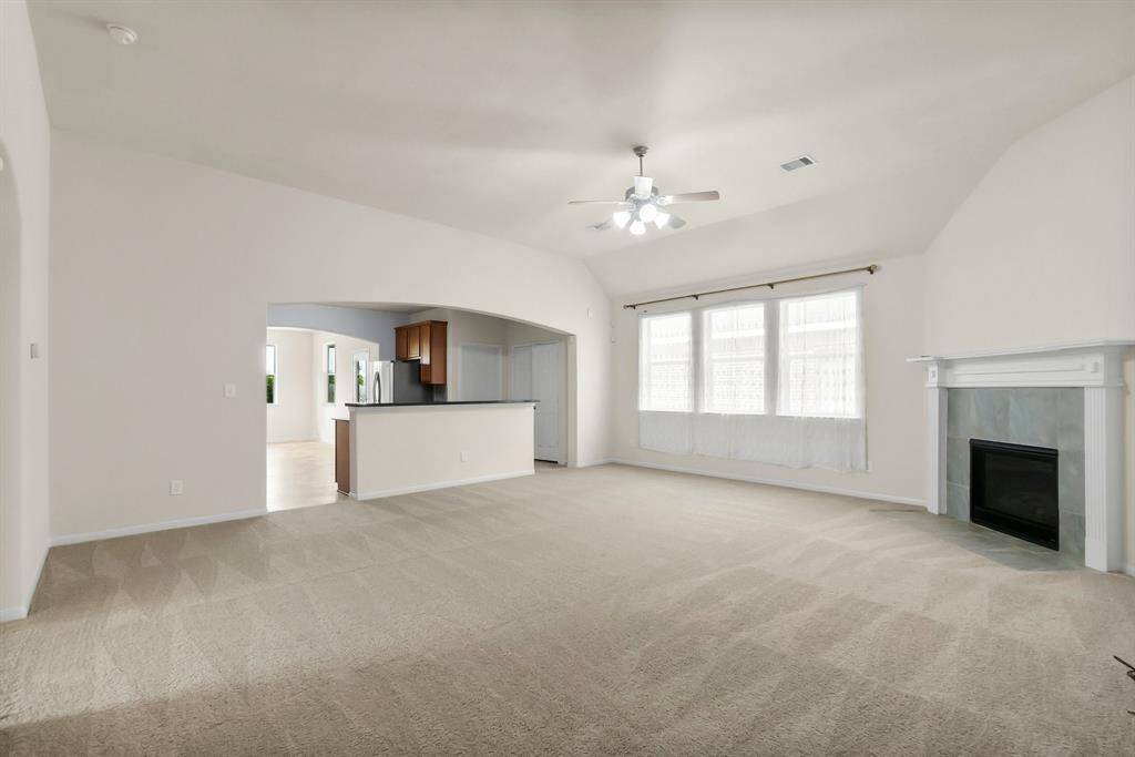 Spacious living room with neutral carpeting, large windows, and a ceiling fan. Features a cozy fireplace and an open view into the kitchen with modern appliances.