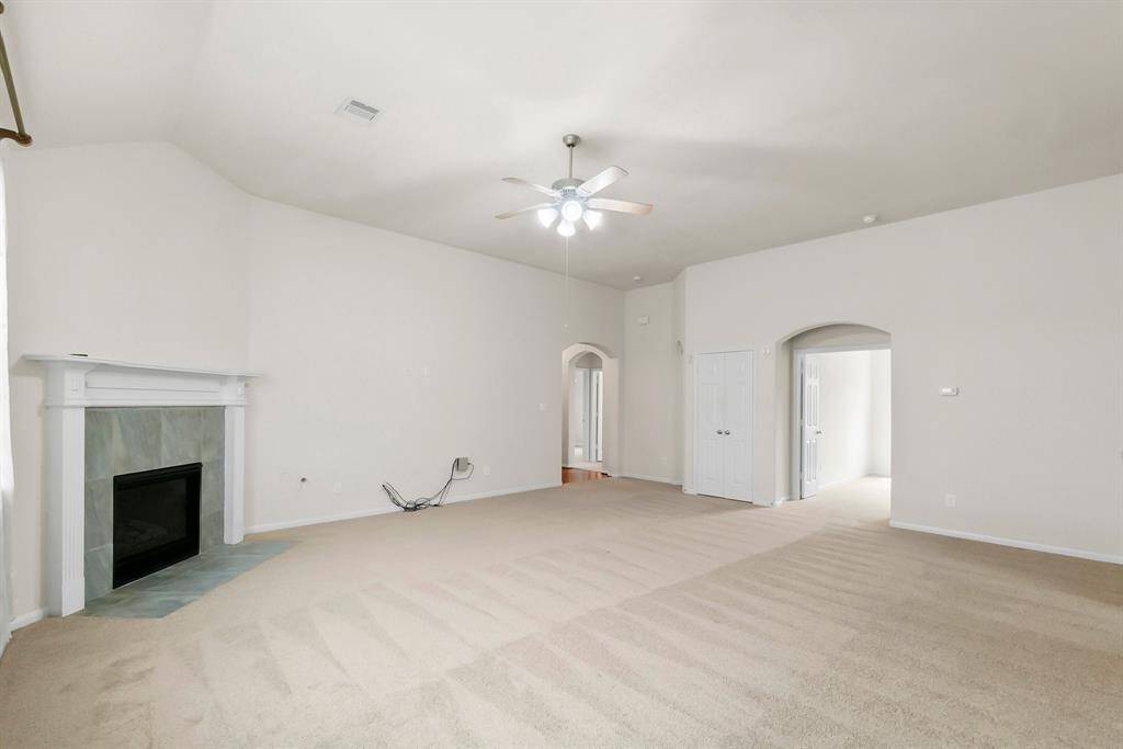 This spacious living room features neutral carpet, a cozy fireplace with a white mantel, and a ceiling fan. The room has high ceilings and leads to other areas through arched doorways, creating an open and inviting atmosphere.