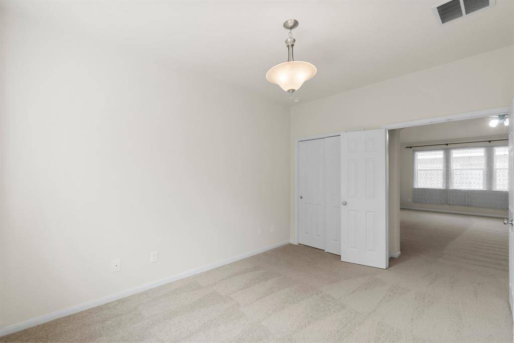This is a bright, neutral-toned room with carpeted flooring, featuring a closet with white double doors and a modern light fixture. It opens to another room with large windows, allowing ample natural light.