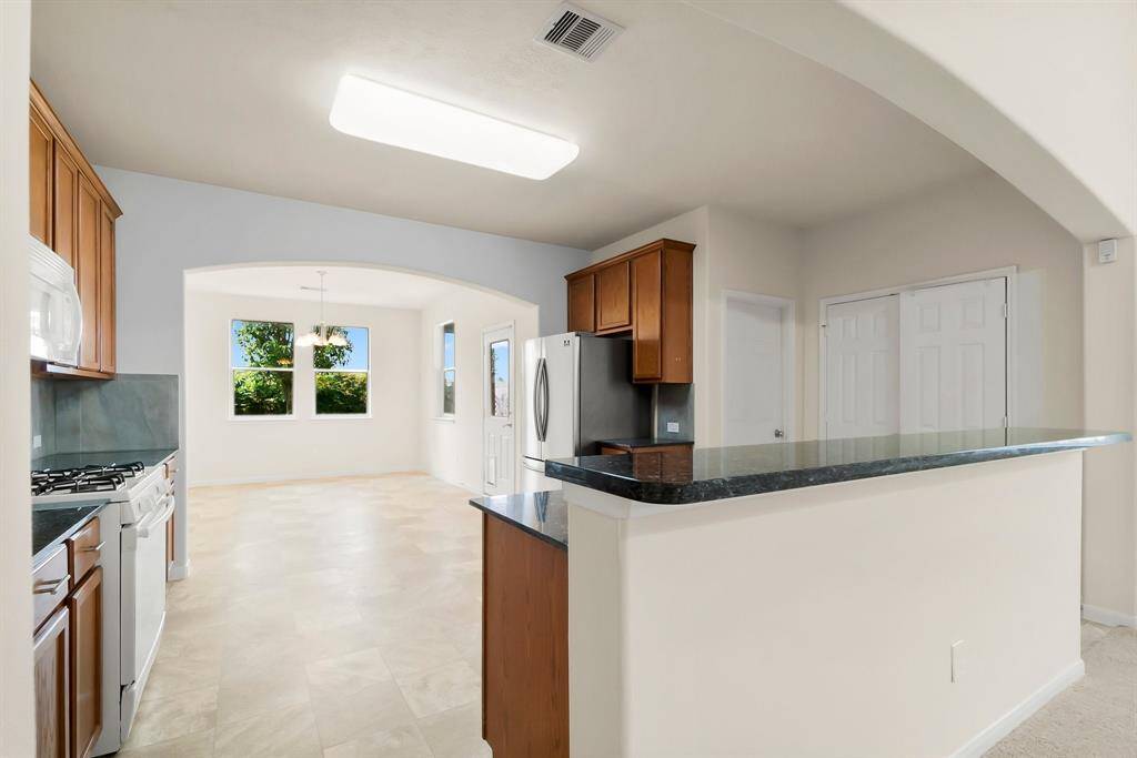 This kitchen features wooden cabinets, black granite countertops, and modern appliances, including a gas stove and stainless steel refrigerator. It opens into a bright dining area with large windows, offering a view of the outdoors.