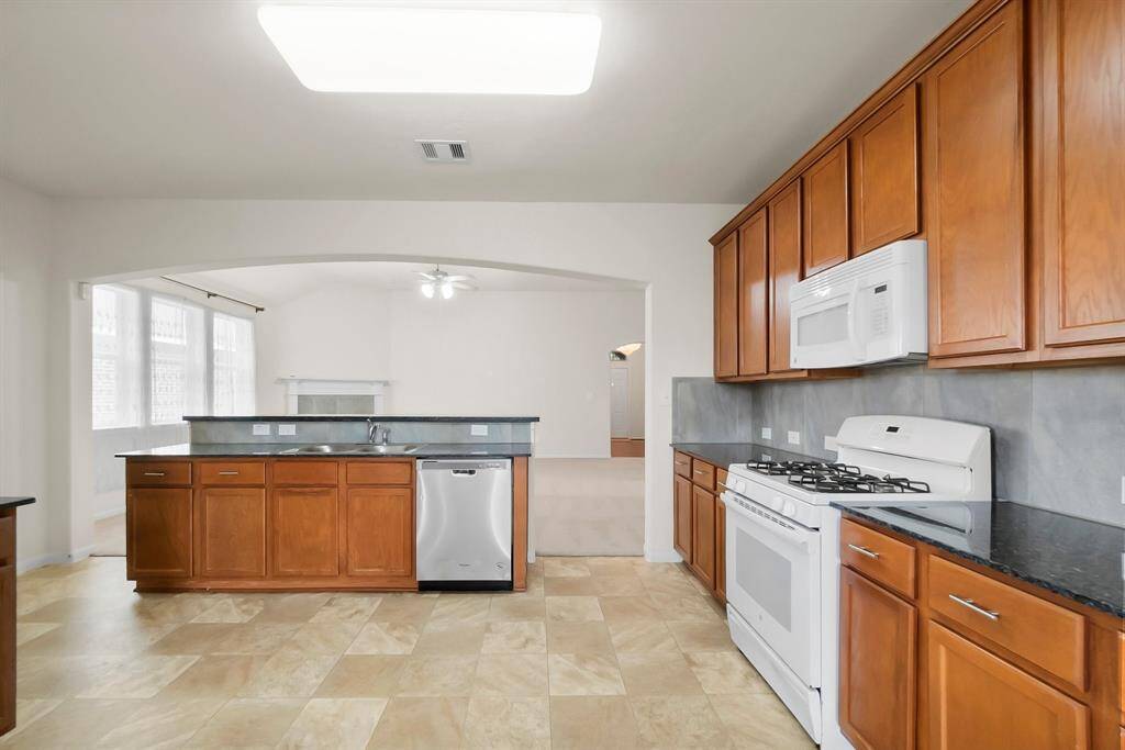 Bright, open kitchen with wood cabinets, a gas stove, and a bar counter with dark countertops. It leads into a dining area with large windows offering a view of greenery, and a side door for easy outdoor access.