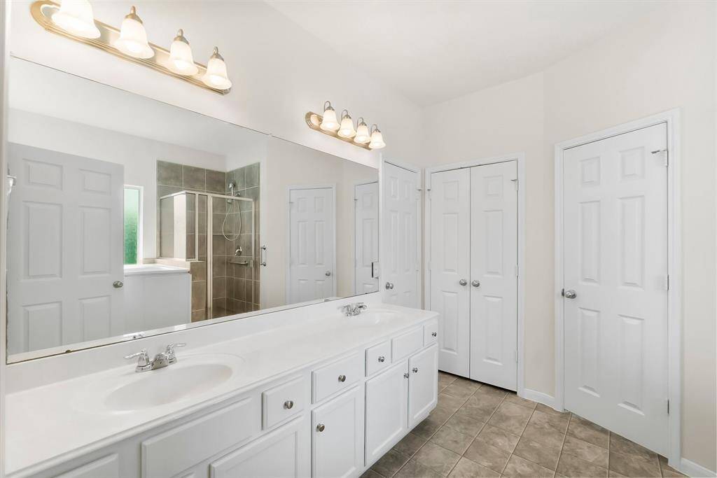 Primary bedroom carpeted with neutral walls and a ceiling fan. It features two doorways: one leading to a bathroom with visible cabinetry, and the other to a hallway. Abundant natural light enters through a window on the left.