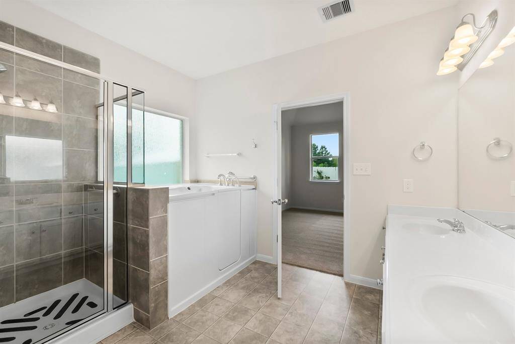 This bright bathroom features a large vanity with ample storage, a spacious mirror, and a walk-in shower with glass doors. The room has neutral tile flooring and two closet doors, offering a clean and functional space.