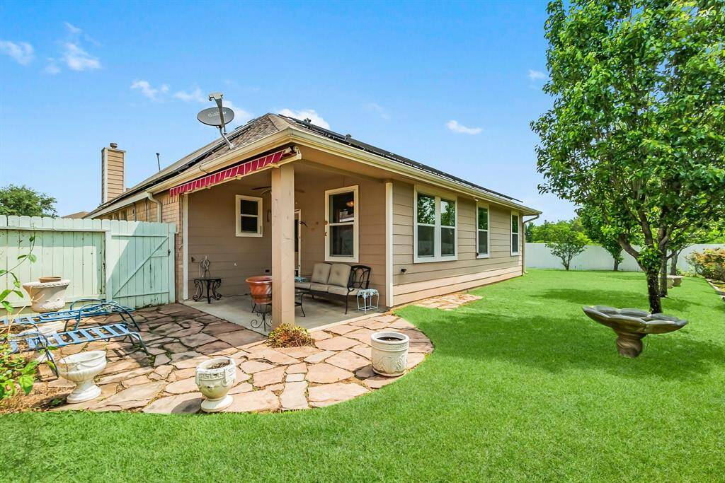 Charming backyard with a covered patio featuring a ceiling fan, perfect for outdoor relaxation. The space includes stone paving, a small seating area, and a well-maintained lawn, all enclosed by a light-colored fence for privacy.