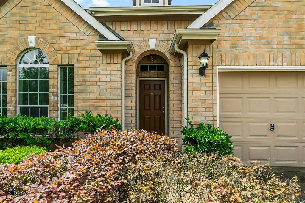 Charming brick exterior with arched windows and a welcoming front door. Features a lantern-style light fixture and well-maintained landscaping, including lush greenery and colorful shrubs. Attached garage for convenience.