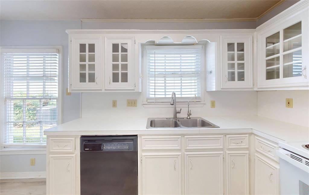 Bright kitchen with white cabinetry, a double sink, and dishwasher. It features large windows, providing ample natural light. Ideal for a fresh and airy cooking space.