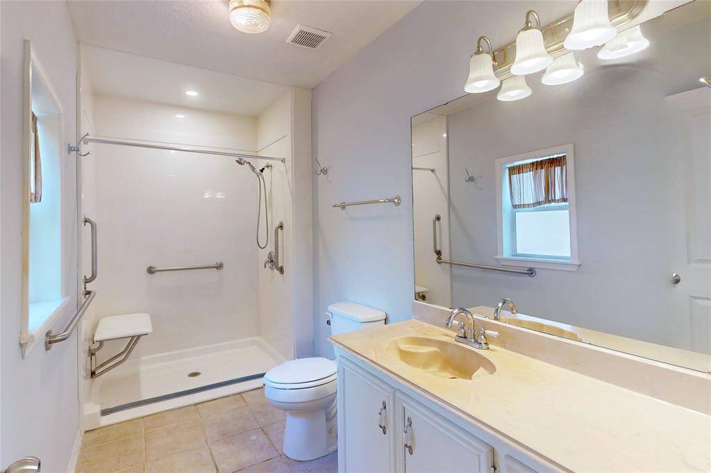 The primary bathroom features a spacious walk-in shower with grab bars and a built-in seat, a large mirror above a single-sink vanity, and neutral tile flooring. Ideal for accessibility and comfort.