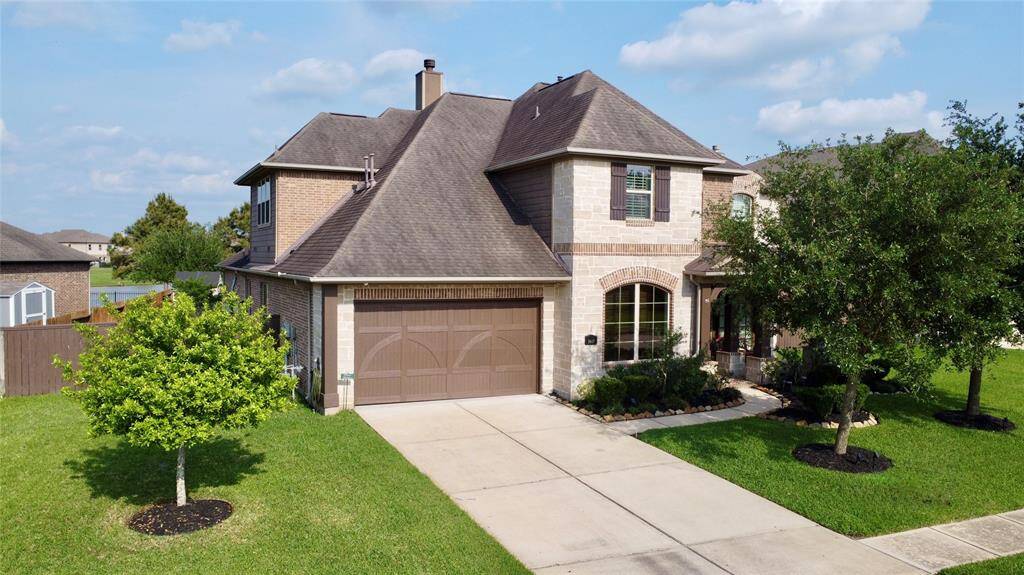 This is a two-story brick home with a gabled roof, featuring a two-car garage and a well-maintained front yard with trees and shrubs. The driveway is spacious, and the exterior has a mix of stone and brick accents, giving it a classic, inviting look.