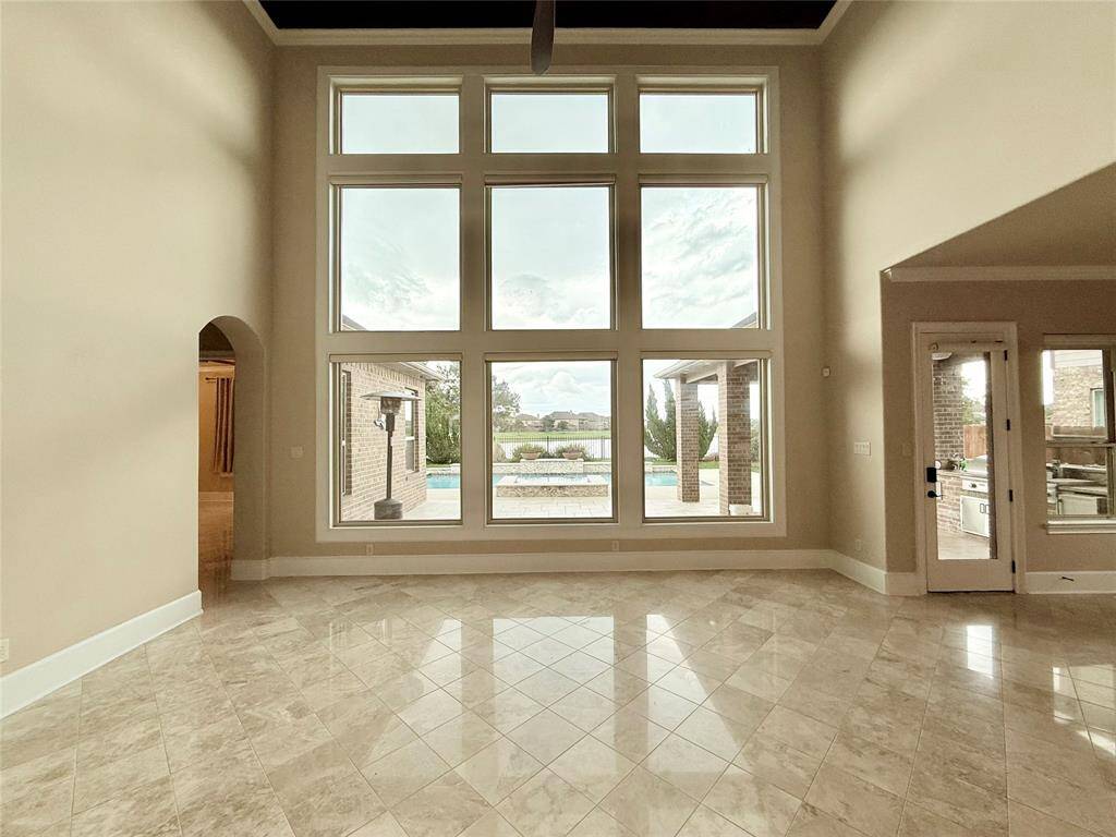 This spacious living area features high ceilings and large windows, offering abundant natural light and a stunning view of the outdoor pool area. The tiled flooring adds elegance, while an adjacent door provides easy access to the patio.
