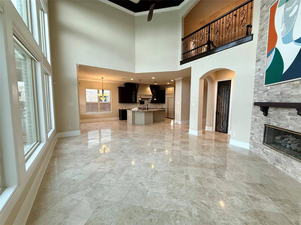 Bright, spacious open-concept living area with high ceilings, large windows, and elegant tile flooring. Features a modern kitchen with dark cabinetry, a stone fireplace, and an upper balcony with wrought iron railing.