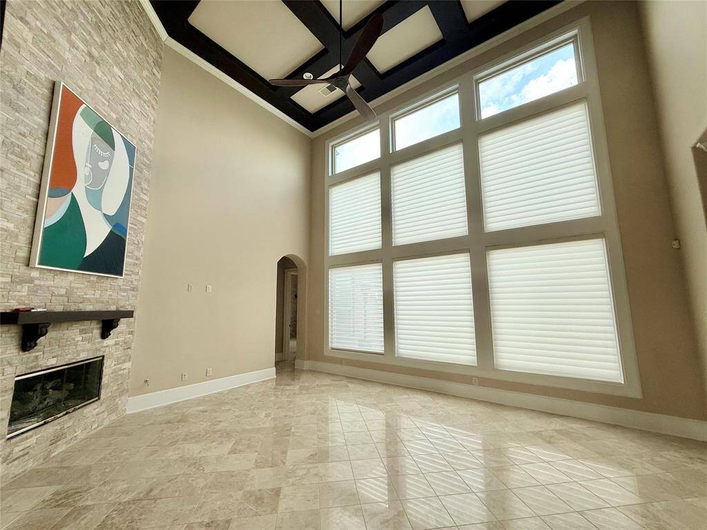 This spacious room features a high ceiling with a modern fan, large windows with blinds for ample natural light, a chic stone accent wall with a fireplace, and elegant tile flooring. Ideal for a bright and welcoming living space.