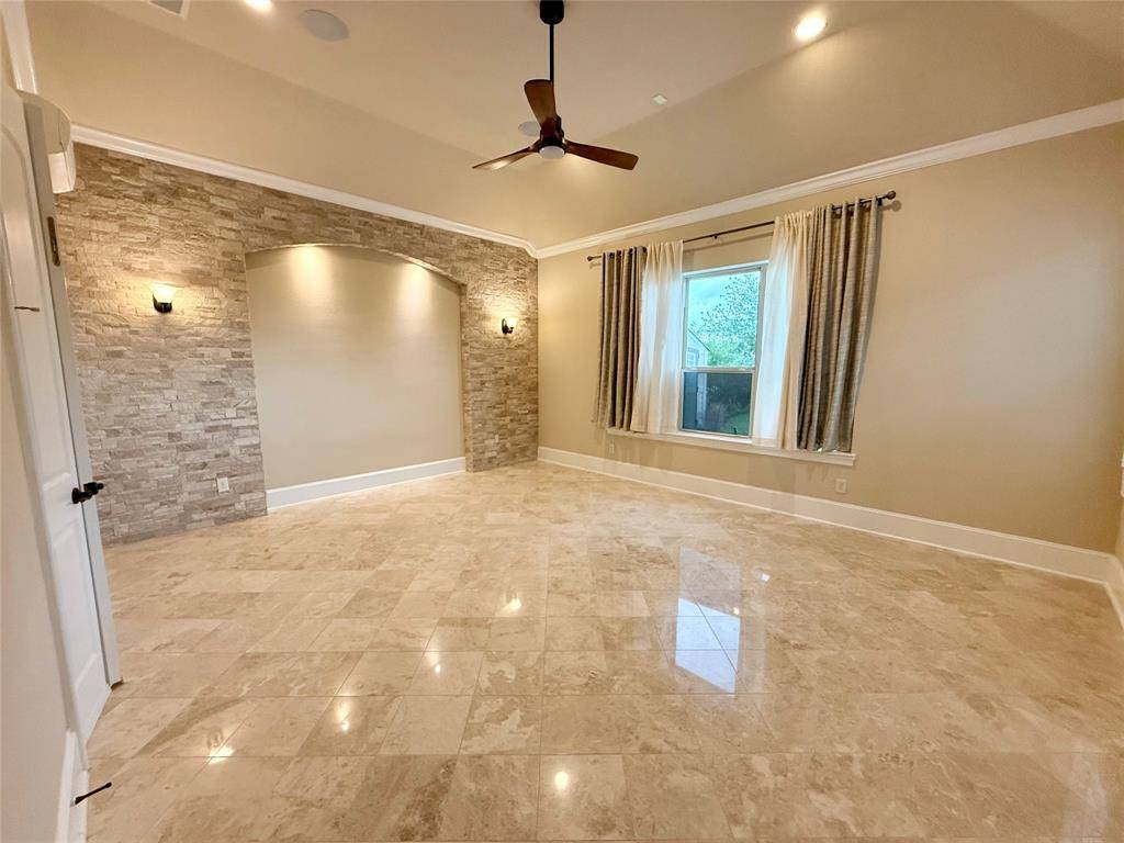 This spacious room features glossy tile flooring, a stone accent wall with built-in lighting, and a large window with curtains. A ceiling fan provides comfort, while the neutral tones offer a modern and elegant feel.