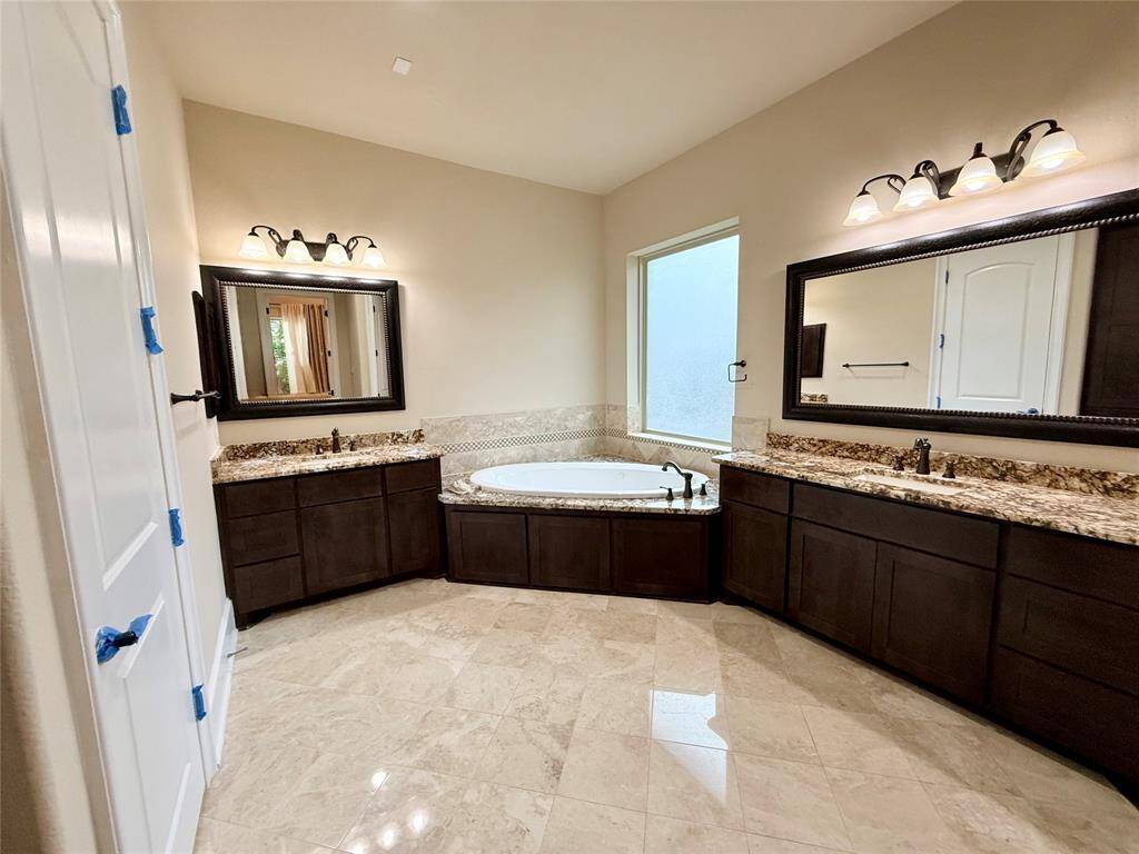 This spacious bathroom features a luxurious corner bathtub, dual vanities with granite countertops, elegant dark wood cabinets, and large mirrors. The natural light from the window enhances the room's warm ambiance.