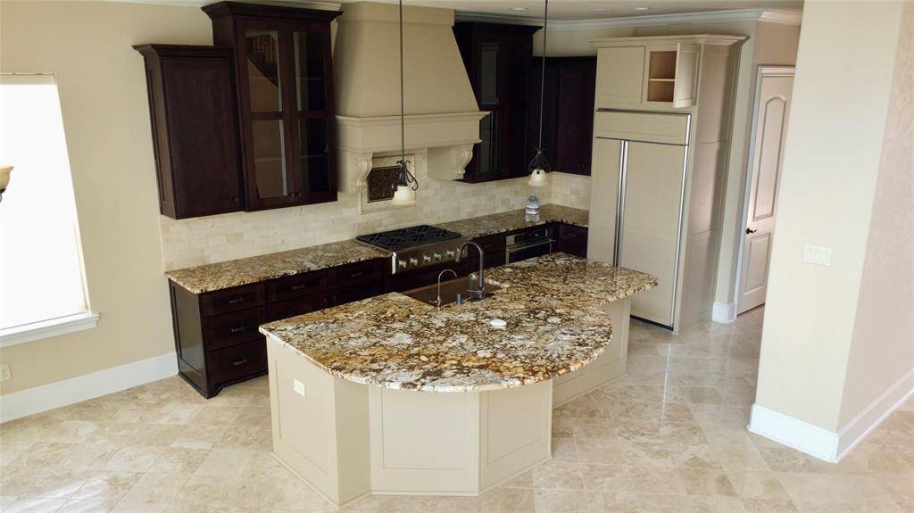 This kitchen features a spacious layout with elegant granite countertops and a central island for added workspace. It includes dark wood cabinetry, stainless steel appliances, and a stylish range hood. The light tile flooring and neutral walls create a warm, inviting atmosphere.