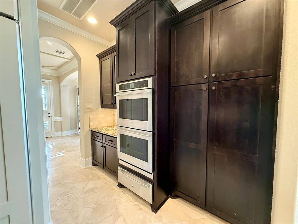 Modern kitchen area with sleek, dark wood cabinets and built-in stainless steel appliances. The space features a light tile floor and a neutral color scheme, providing a clean and sophisticated look. An arched doorway leads to an adjacent area, adding architectural interest.