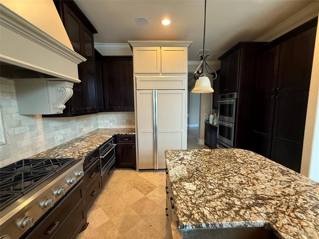 This kitchen features dark wood cabinetry, a large gas stove, and stone countertops with a striking pattern. It includes double ovens, a built-in fridge, and elegant pendant lighting, creating a luxurious and functional cooking space.