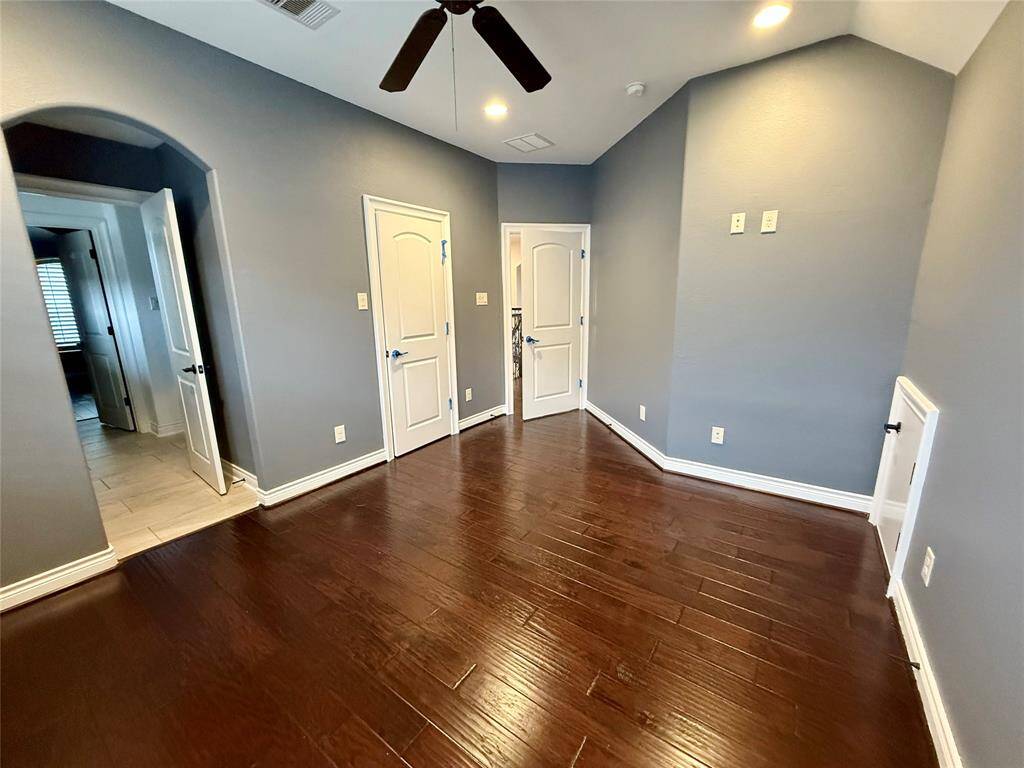 This room features rich hardwood flooring, soft gray walls, and a ceiling fan. It includes multiple doors and an arched entryway leading to a tiled area, offering a modern and versatile space.