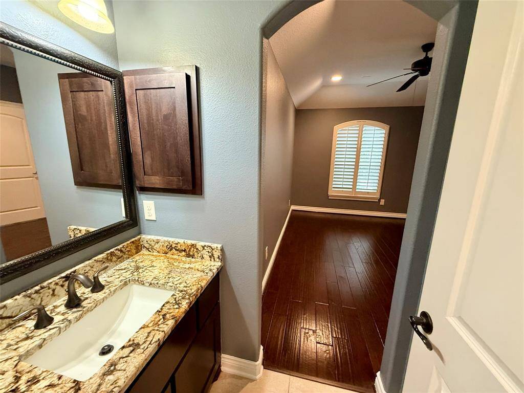 Modern bathroom with a granite countertop and dark wood cabinetry. It opens into a room with hardwood floors, a ceiling fan, and a window with shutters, creating a cozy and stylish living space.
