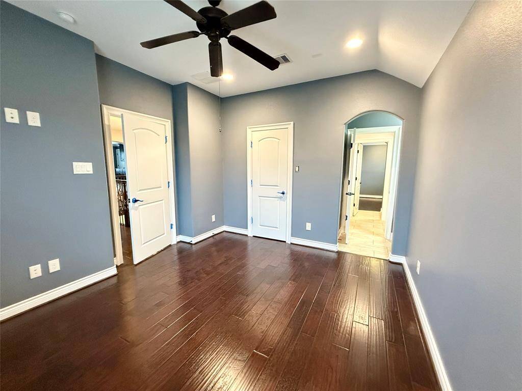 This room features rich hardwood floors, soft blue-gray walls, and a ceiling fan. It includes multiple doors leading to adjacent rooms, offering a spacious and connected feel.
