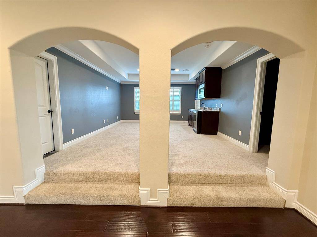Spacious room with carpeted area divided by an archway. The space features dark hardwood floors leading to a cozy area with gray walls, a mini bar with cabinetry, and natural light from a window. Ideal for entertaining space.