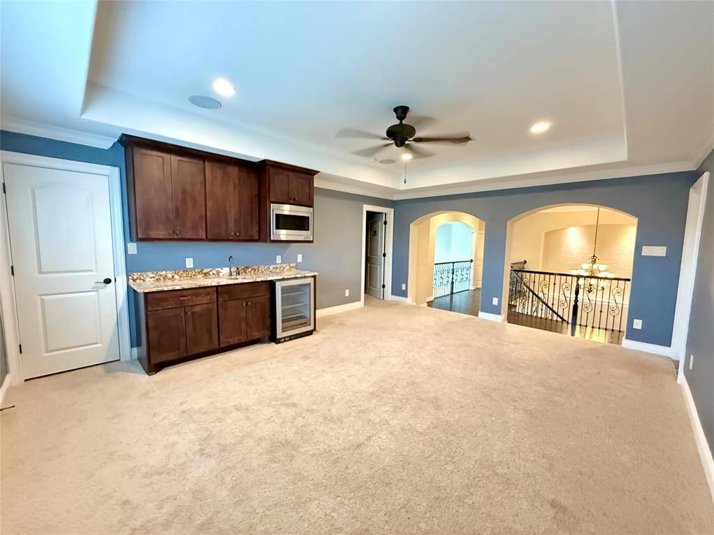 This spacious room features a wet bar with granite countertops, microwave, and mini fridge. It has plush carpeting, a ceiling fan, and overlooks a staircase through elegant archways, offering a versatile living area.