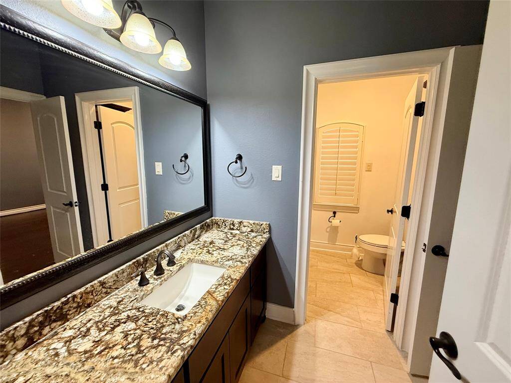Modern bathroom with a granite countertop and a large mirror. The room features elegant lighting and a separate space for the toilet with a shuttered window. The walls are painted a soft blue-gray, creating a serene atmosphere.
