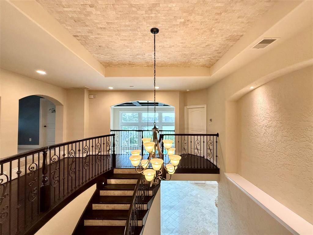 Spacious and elegant foyer with a grand staircase and wrought iron railing. The ceiling features intricate detailing and a stylish chandelier. Large windows provide natural light, enhancing the open, airy feel.