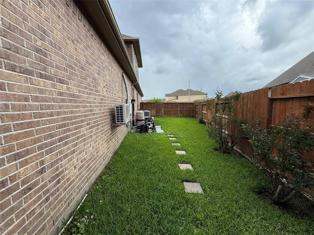 Narrow side yard with a brick exterior wall, green grass, stepping stones, and a wooden fence. There are some small plants along the fence, and air conditioning units are attached to the house.
