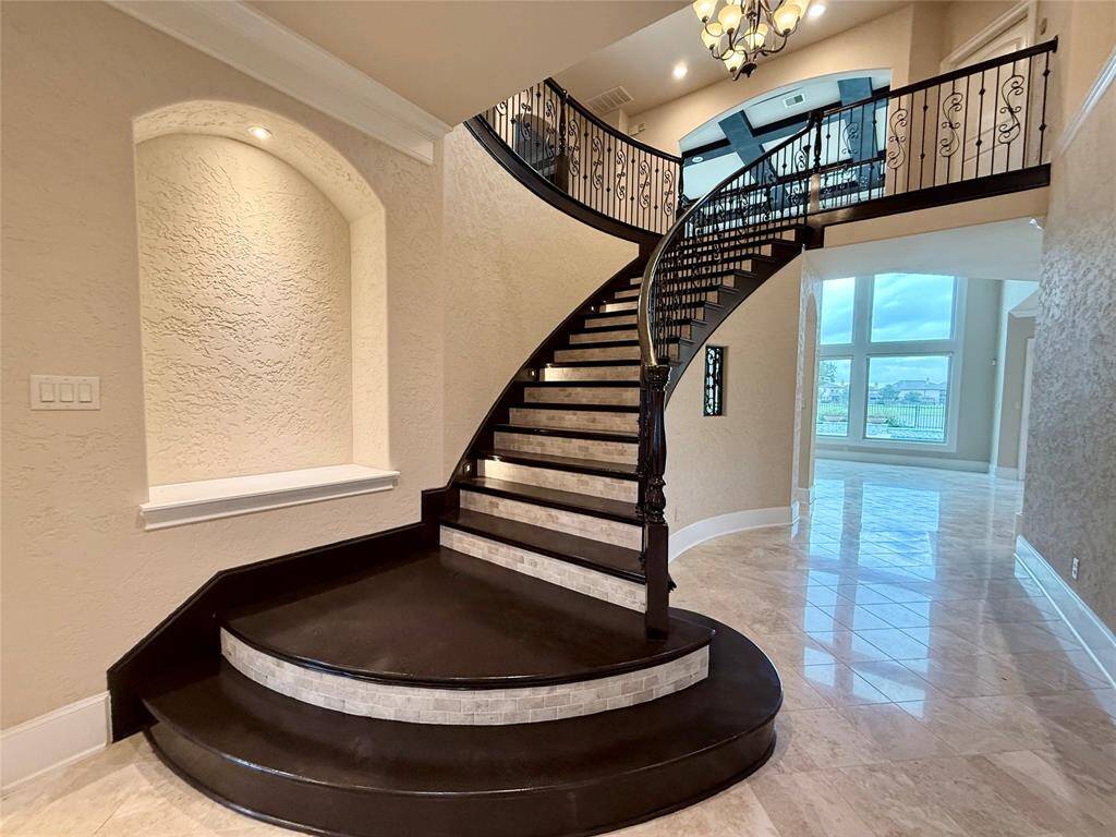 After opening the door, you'll find an elegant entryway with a grand, curved staircase featuring wrought-iron railings and dark wood steps. The space is highlighted by a chandelier and textured walls, leading to a bright, open area with large windows offering scenic views.