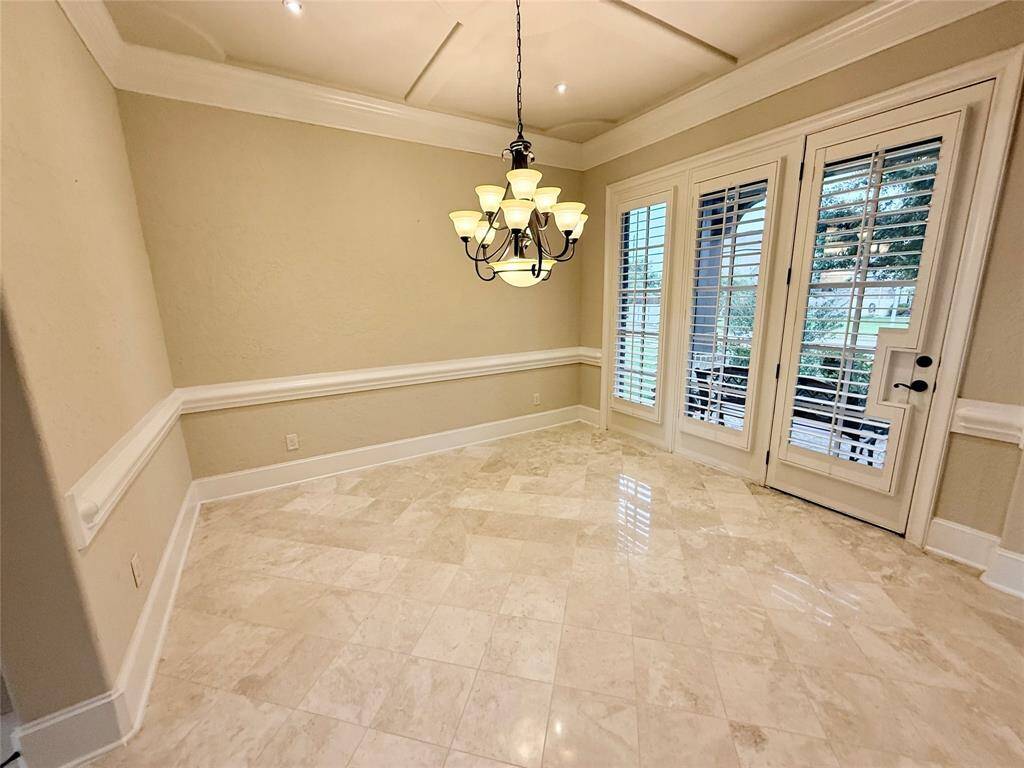 This elegant dining room features polished marble floors, a stylish chandelier, and French doors with plantation shutters, offering a bright and sophisticated space.