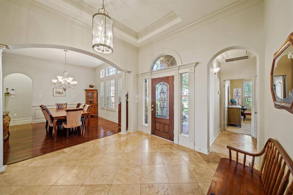 This inviting entryway features a grand wooden door with decorative glass panels, leading to a spacious foyer with elegant tile flooring. Arched doorways open into a dining area with rich wood flooring and a stylish chandelier. The space is bright and welcoming, perfect for greeting guests.