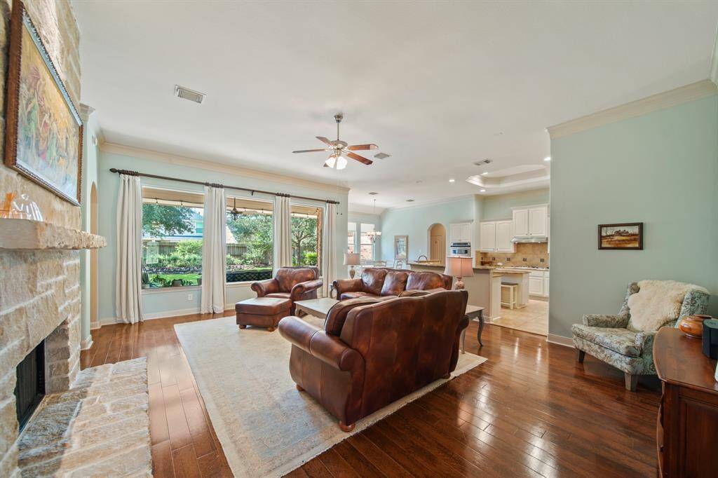 This spacious living area features hardwood floors, a cozy stone fireplace, and large windows offering plenty of natural light. The open layout flows into a modern kitchen with white cabinetry, perfect for entertaining.